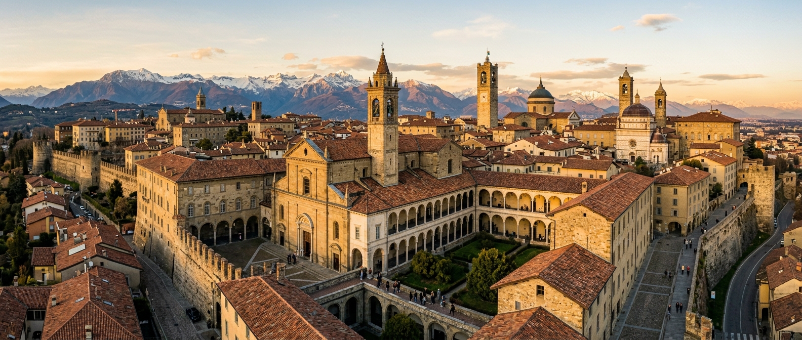 Panoramic view of University of Bergamo Humanities campus in the medieval Upper City (Città Alta), historic stone buildings with arched cloisters, bell towers visible against the backdrop of the Bergamo Alps, warm golden hour light